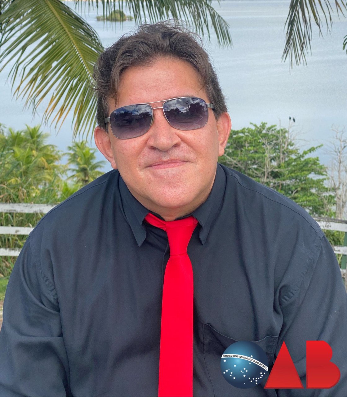 Foto de Melhym Quemel. Homem com pele clara, cabelos escuros, usando óculos escuros estilo aviador, camisa social preta e gravata vermelha. Ao fundo, palmeiras e um corpo d'água. No canto inferior direito, há a marca d'água com o logotipo da OAB (esfera azul com o Cruzeiro do Sul e as letras OAB em vermelho).
